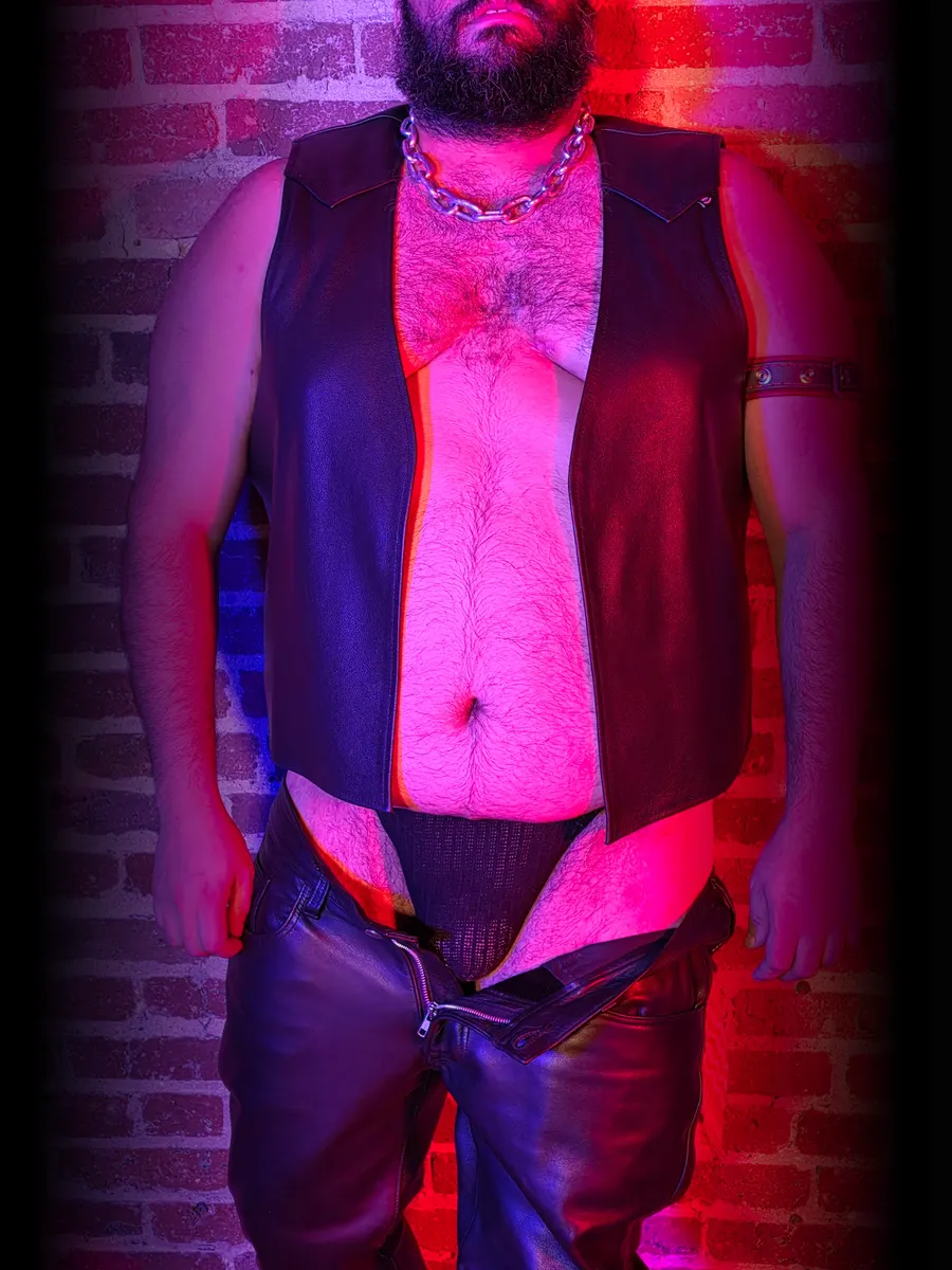 A bearded dude in an open leather vest and leather pants over a jock, lit in red and blue